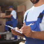 Renting vs. Buying Packing Supplies: Tips for Minimizing Packing Material Costs 2 Two moving workers in blue overalls are loading boxes into a moving truck. One worker is holding a clipboard and pen, taking notes, while the other is arranging boxes inside the truck. The scene depicts a busy moving day, emphasizing teamwork and organization in the moving process.