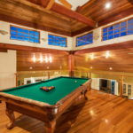 Renting vs. Buying Packing Supplies: Tips for Minimizing Packing Material Costs 5 Interior view of a stylish game room featuring a green felt billiard table, wooden flooring, and large windows that provide natural light. The room showcases a warm wooden ceiling and modern lighting, creating an inviting atmosphere for leisure and entertainment.