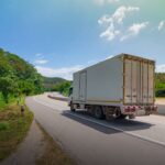 How Much Does it Cost to Get People to Help You Move? 2 A large delivery truck driving on a winding road surrounded by lush greenery and clear blue skies, showcasing transportation in a rural landscape.