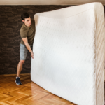 The Benefits of Using Picture Moving Boxes When Packing 5 Young man lifting a large mattress in a modern bedroom with wood flooring and a stone accent wall, showcasing the process of moving or rearranging furniture.