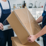 How Much Are Movers Help Per Hour in NYC? 5 Two movers in blue overalls are wrapping a cardboard box with plastic wrap in a tidy, modern kitchen environment. Several unpacked boxes are stacked nearby, indicating an ongoing moving process.