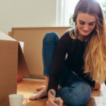 Packing a Mattress for Moving: Everything You Need To Know 6 Young woman sitting on the floor, surrounded by cardboard boxes, writing notes with a pen. A coffee cup is nearby, indicating a cozy moving day atmosphere. Natural light filters through a window, enhancing the warm and productive environment.