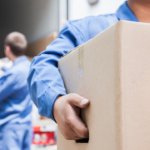 How to Save Money on Local Moving Services 5 Workers in blue uniforms handling cardboard boxes during a moving process, with one individual prominently carrying a box while others load additional boxes into a vehicle.