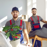 Why Would I Want to Use White Glove Moving? 4 Two smiling movers in uniforms carrying a potted plant and a modern chair in a bright, airy living room. The scene showcases professional moving services in a residential setting.