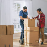 Renting vs. Buying Packing Supplies: Tips for Minimizing Packing Material Costs 4 Two men collaborating on moving boxes in a spacious, well-lit room. One man is checking off items on a list while the other stands next to a hand truck loaded with cardboard boxes. The room features large windows and a couch covered in plastic.