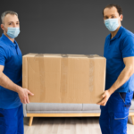 Long Distance Moving: Why Hiring Professionals Is Worth The Investment 3 Two delivery workers in blue uniforms and face masks are holding a large cardboard box in a modern living room. The background features a gray wall and a sofa, emphasizing a clean and organized environment suitable for moving services.