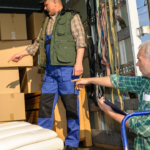 Your Guide to Long Distance Moving in NYC: Stress Less, Move More 6 Two men are working inside a moving truck filled with stacked cardboard boxes. One man, wearing a green vest and blue pants, is pointing towards a white sofa, while the other man, dressed in a plaid shirt, is holding a clipboard and observing the situation. The image captures the teamwork involved in loading and organizing items during a move.