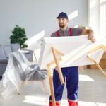 Why Would I Want to Use White Glove Moving? 3 A moving professional in a cap and uniform holds a white table upside down in a bright, modern living room. The space features a gray sofa, a potted plant, and cardboard boxes, indicating a recent move or renovation. Sunlight streams through large windows, enhancing the fresh, organized atmosphere.