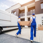 Long Distance Moving: Why Hiring Professionals Is Worth The Investment 4 Two movers in blue overalls carrying a cardboard box from a delivery van to a modern residential building with a paved driveway.