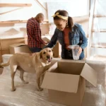 Long Distance Moving: Why Hiring Professionals Is Worth The Investment 2 A woman smiles while petting a Labrador dog next to a cardboard box during a home moving process, with a man in the background organizing boxes. The scene shows a cozy, unpacking environment filled with packing materials and wooden furniture.