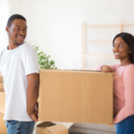 How Do White Glove Movers Work 3 A young couple smiles while carrying a cardboard box in a bright, modern living room filled with moving boxes, symbolizing the excitement of moving into a new home.